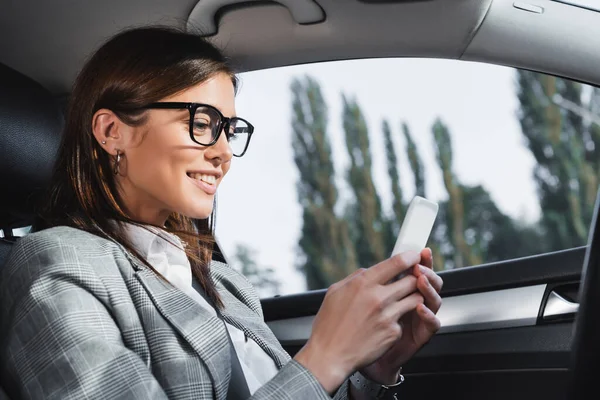 Mulher de negócios sorridente em óculos mensagens no telefone celular enquanto sentado no carro — Fotografia de Stock