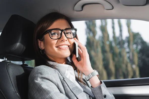 Happy businesswoman in eyeglasses looking at camera while talking on smartphone in car — Stock Photo