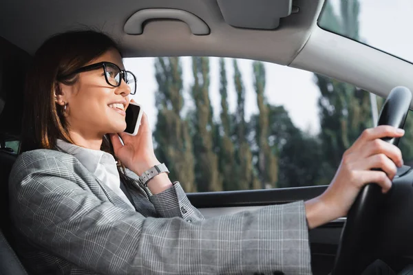 Cheerful businesswoman in eyeglasses driving car and talking on smartphone — Stock Photo