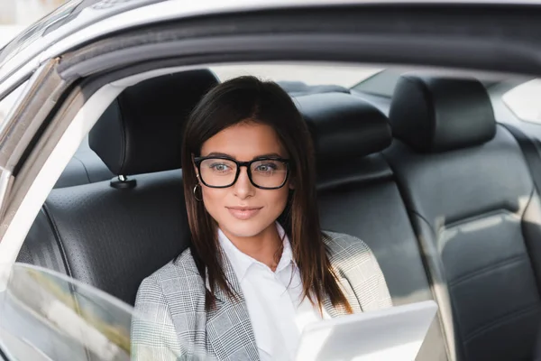Smiling businesswoman with digital tablet looking out from open window while riding in car on blurred foreground — Stock Photo