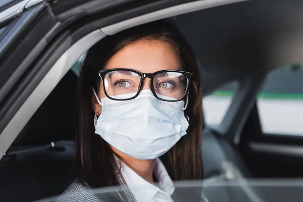 Young businesswoman in eyeglasses and medical mask looking out from open window while riding in car — Stock Photo