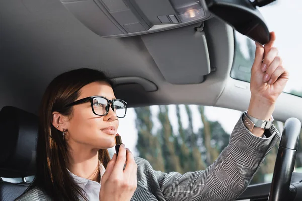 Elegante mujer de negocios sosteniendo lápiz labial mientras se ajusta espejo retrovisor en el coche - foto de stock