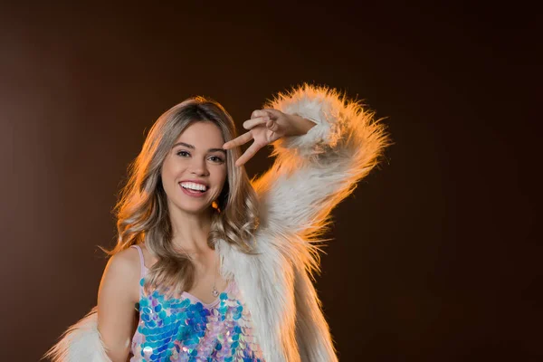 Pleased blonde woman in faux fur jacket showing peace sign on black — Stock Photo