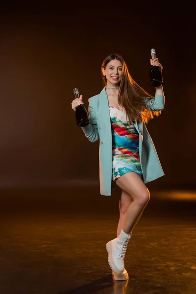 Comprimento total de mulher morena alegre na jaqueta segurando garrafas com champanhe no preto — Fotografia de Stock