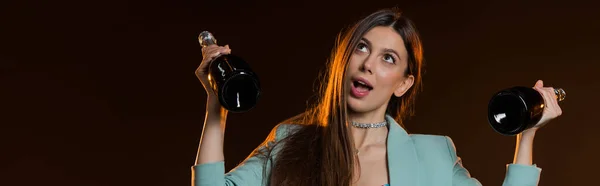 Surprised brunette woman in jacket holding bottles with champagne on black, banner — Stock Photo