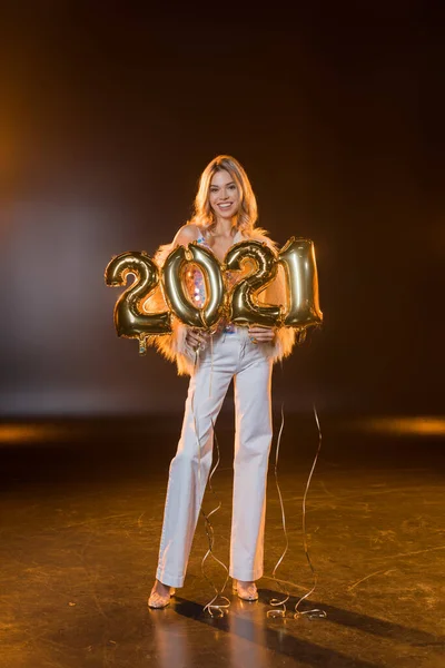 Full length of cheerful blonde woman smiling and holding 2021 numbers balloons on black — Stock Photo