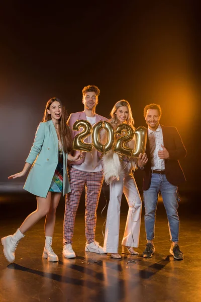 Full length of happy multicultural friends holding 2021 numbers balloons on black — Stock Photo