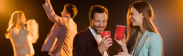 Счастливая женщина и африканский американец мужчина держит пластиковые чашки рядом с друзьями во время вечеринки на черном, баннер — стоковое фото