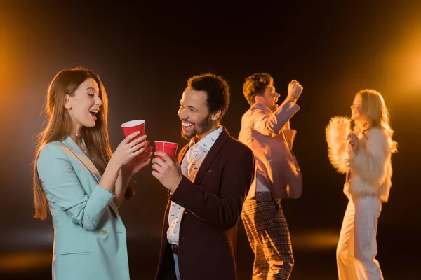 Cheerful woman and african american man holding plastic cups near friends dancing during party on black — Stock Photo