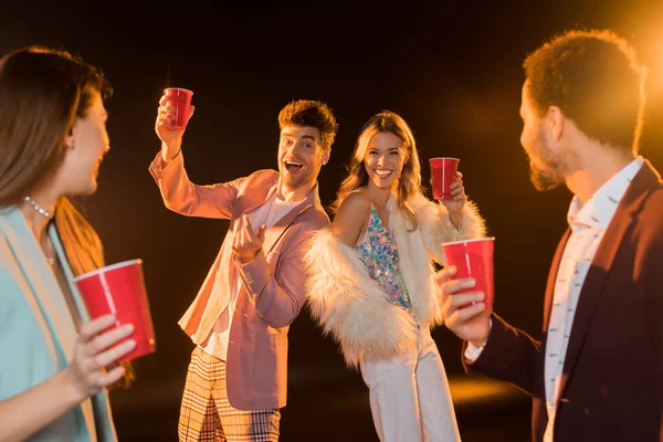 Excité homme et femme tenant des tasses en plastique près d'amis multiculturels sur le premier plan flou sur noir — Photo de stock