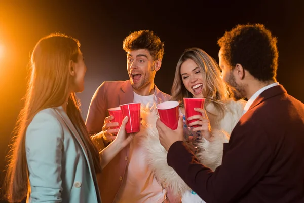 Group of excited multicultural friends toasting plastic cups during party on black — Stock Photo