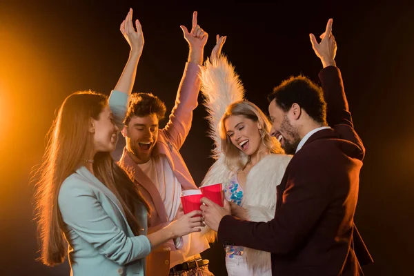 Group of excited multicultural friends toasting plastic cups and rejoicing during party on black — Stock Photo