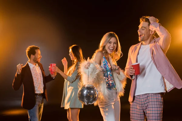 Mulher loira feliz segurando bola de discoteca perto do homem com copo de plástico e amigos multiculturais no fundo borrado no preto — Fotografia de Stock