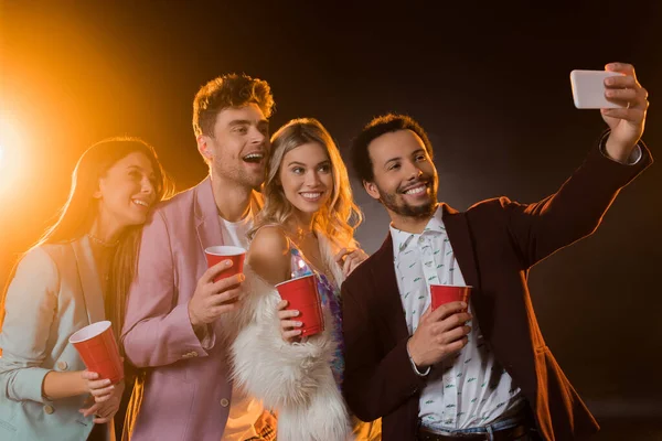 Group of cheerful multicultural friends taking selfie while holding plastic cups on black — Stock Photo