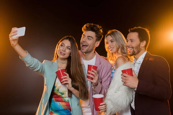 Group of happy multicultural friends taking selfie while holding plastic cups on black — Stock Photo