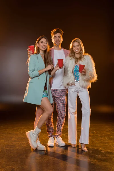 Comprimento total de homem alegre e mulheres segurando copos de plástico durante a festa em preto — Fotografia de Stock