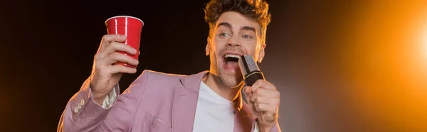 Man singing while holding microphone and plastic cup on black, banner — Stock Photo