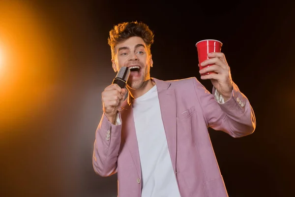 Hombre cantando mientras sostiene el micrófono y la taza de plástico en negro - foto de stock