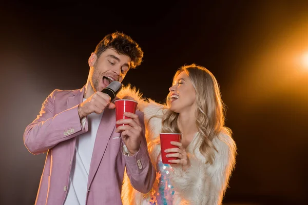 Homme chantant tout en tenant microphone près de la femme avec tasse en plastique sur noir — Photo de stock