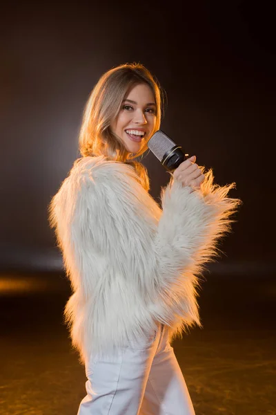 Mujer rubia feliz cantando mientras sostiene el micrófono en negro - foto de stock