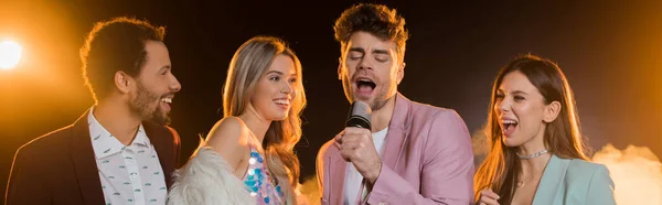 Man singing while in microphone near multicultural friends on black, banner — Stock Photo