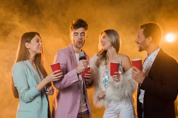 Hombre cantando mientras en el micrófono cerca de amigos multiculturales con copas de plástico en negro con humo - foto de stock