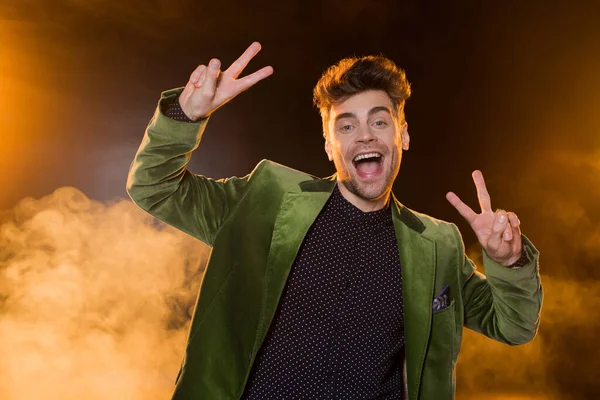 Hombre excitado en chaqueta verde mostrando signo de paz en negro con humo - foto de stock