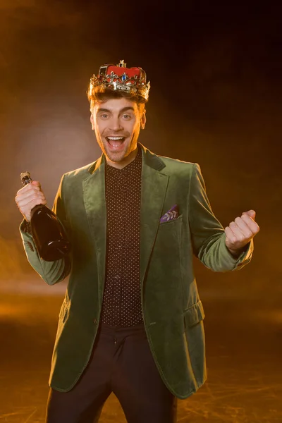 Excited man in green blazer and crown holding bottle of champagne on black — Stock Photo