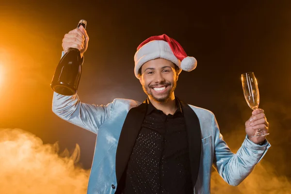 Aufgeregter afrikanisch-amerikanischer Mann mit Weihnachtsmütze, der eine Flasche Champagner und Glas auf schwarz mit Rauch hält — Stockfoto