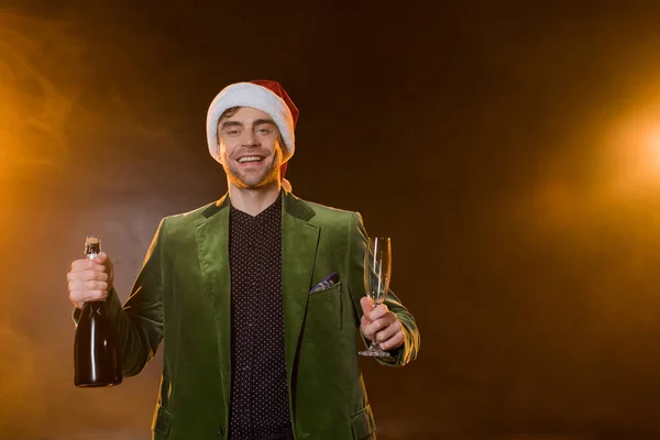 Cheerful man santa hat holding bottles of champagne and glasses on black — Stock Photo