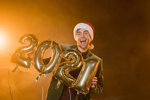 Aufgeregter Mann mit Weihnachtsmütze hält Luftballons mit den Zahlen 2021 auf schwarz — Stockfoto