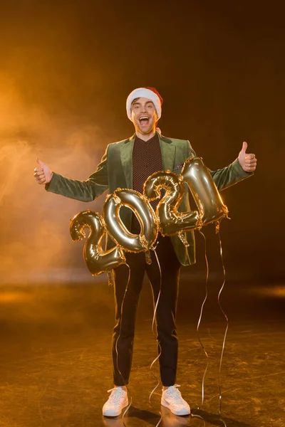 Pleine longueur de l'homme excité dans le chapeau de Père Noël montrant pouces vers le haut près de ballons avec 2021 numéros sur noir — Photo de stock