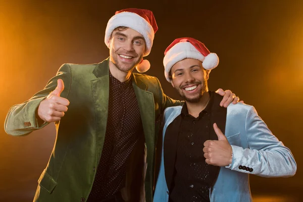 Felici uomini interrazziale in cappelli di Babbo Natale mostrando pollici su sul nero — Foto stock