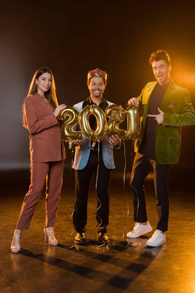 Comprimento total de homem americano africano feliz na coroa segurando balões com 2021 números perto de amigos em preto — Fotografia de Stock