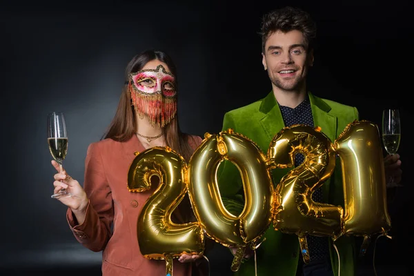 Felice uomo che tiene palloncini con 2021 numeri vicino alla donna in maschera di carnevale con un bicchiere di champagne sul nero — Foto stock