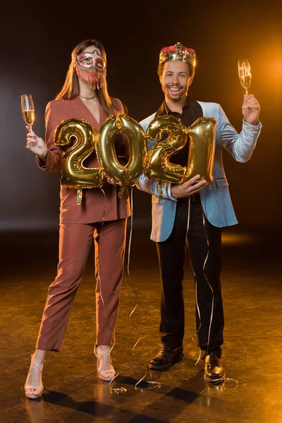 Comprimento total de homem americano africano feliz em coroa segurando balões com 2021 números perto de mulher em máscara de carnaval com copo de champanhe em preto — Fotografia de Stock