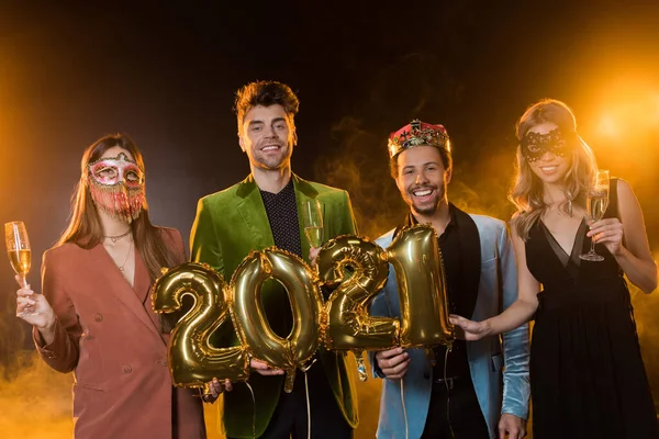 Fröhliche multikulturelle Männer mit Luftballons mit Zahlen 2021 neben Frauen mit Gläsern Champagner auf Schwarz — Stockfoto