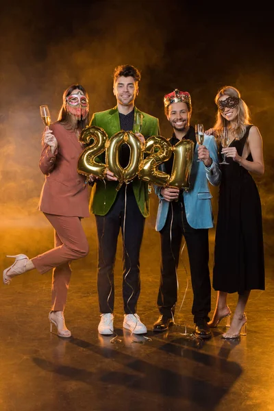 Volle Länge fröhlicher multikultureller Männer mit Luftballons mit Zahlen 2021 neben Frauen mit Gläsern Champagner auf Schwarz — Stockfoto