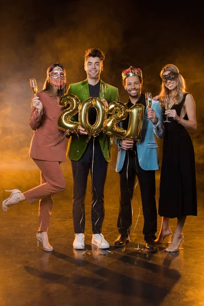 Full length of happy multicultural men holding balloons with 2021 numbers near women with glasses of champagne on black — Stock Photo