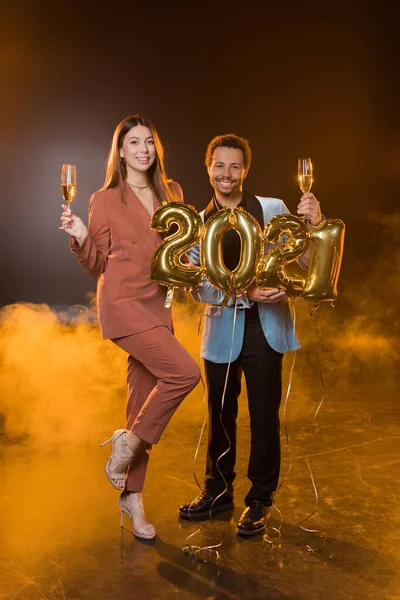 Longitud completa de feliz pareja multicultural sosteniendo copas de champán cerca de globos con 2021 números en negro - foto de stock
