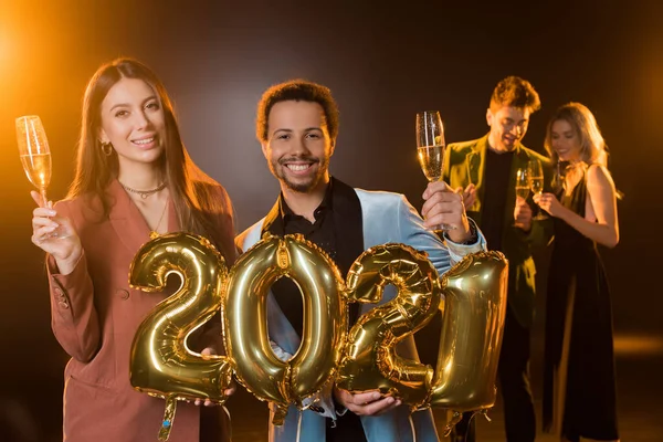 Feliz casal multicultural segurando copos de champanhe e balões com 2021 números perto de amigos no fundo borrado em preto — Fotografia de Stock
