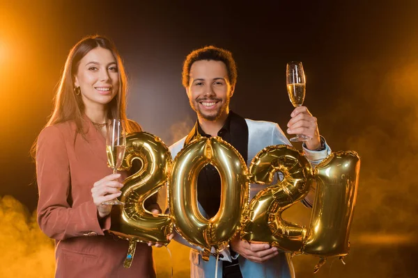 Happy multicultural couple holding glasses of champagne near balloons with 2021 numbers on black — Stock Photo