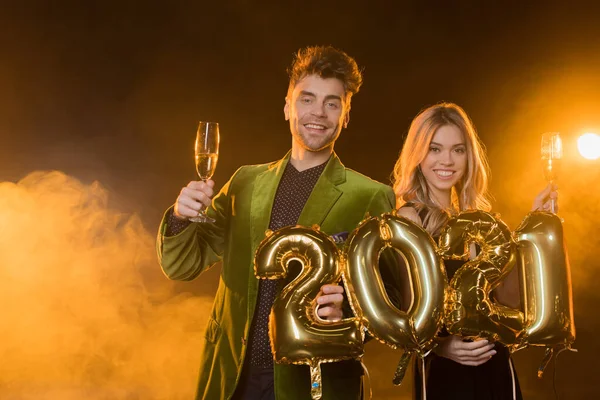 Homem feliz segurando balões com 2021 números perto de mulher com copo de champanhe em preto — Fotografia de Stock