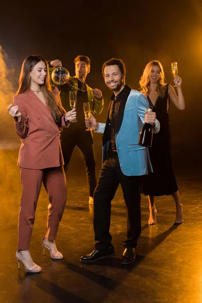 Full length of happy multicultural couple holding glasses of champagne and bottle near friends on blurred and black background — Stock Photo