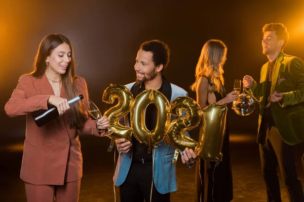 Happy woman pouring champagne in glass near african american man and friends on blurred and black background — Stock Photo