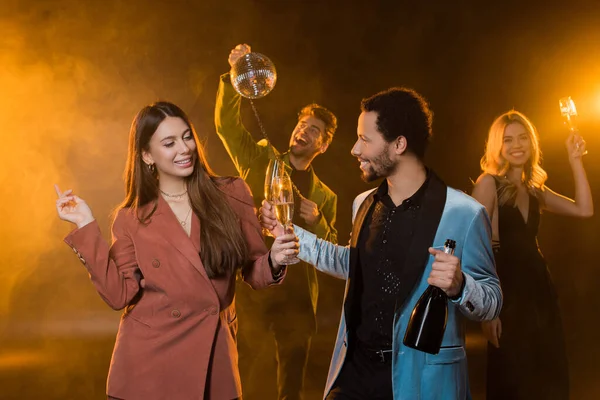 Щаслива мультикультурна пара тримає келихи шампанського та пляшки біля веселих друзів на розмитому та чорному тлі — стокове фото