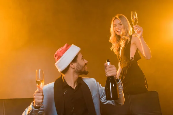 Homme afro-américain joyeux tenant bouteille de champagne et de verre près de femme blonde sur noir avec de la fumée — Photo de stock