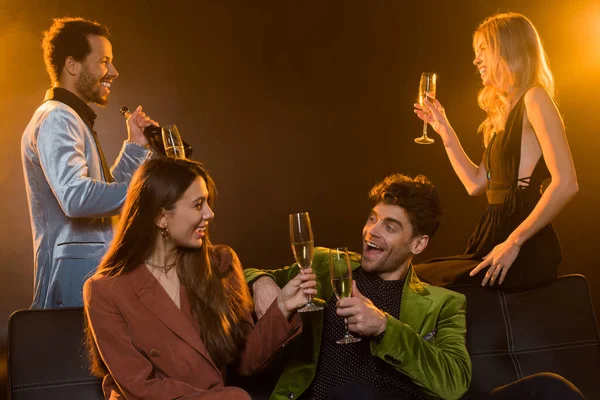 Felice uomo e donna che tiene i bicchieri con champagne mentre si siede sul divano vicino agli amici sul nero — Foto stock