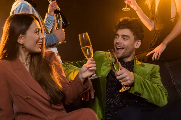 Happy couple holding glasses of champagne while sitting on sofa near friends on black — Stock Photo