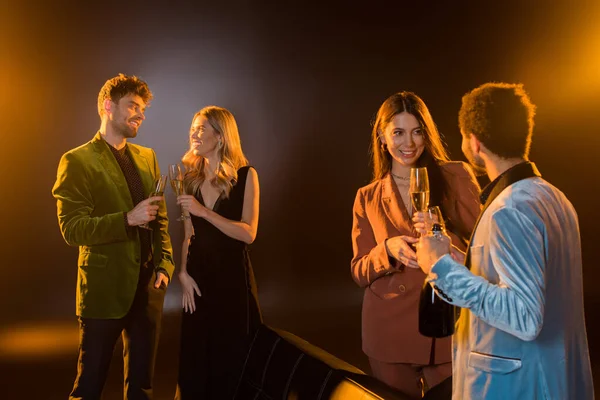 Fröhliche multikulturelle Freunde stoßen mit Champagner auf Glas an und lächeln auf schwarzem Hintergrund — Stockfoto